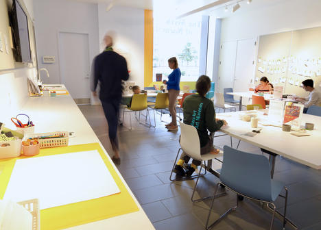 A group of people sitting and standing in a room with arts and crafts tools on a white table.