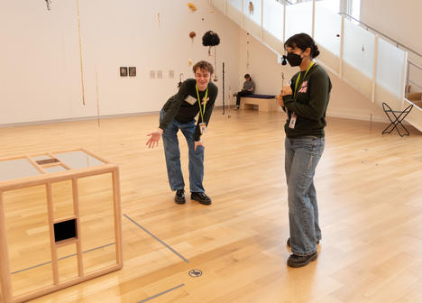 One person standing with a mask beside another person pointing at a mirrored cube.