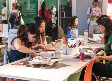 A group of people sitting at tables, engaged in arts and crafts with magazines and scissors. The setting is lively, with colorful chairs and a creative atmosphere.