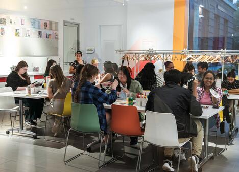 A diverse group of people engage in crafts at tables in a bright, modern room. The atmosphere is lively, with colorful chairs and creative materials.