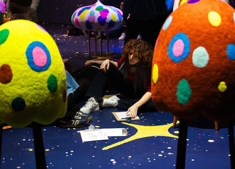 A person with curly hair sits on a starry floor, surrounded by colorful, spotted sculptures resembling mushrooms. They are drawing on a clipboard, creating a thoughtful ambiance.