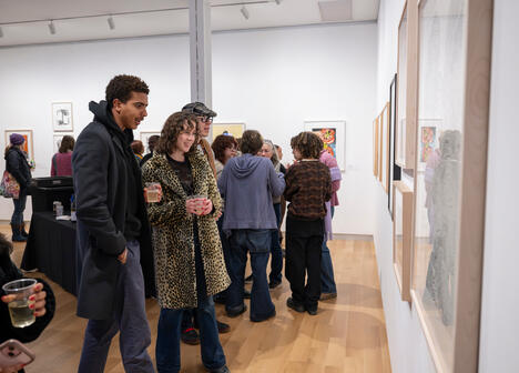 A lively art gallery scene with people admiring framed artworks on white walls. Visitors, casually dressed, hold drinks, showing engagement and interest.
