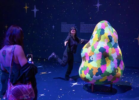 A person poses playfully beside a colorful, abstract sculpture in a dark, starry-themed room. Another person photographs with a phone, creating a joyful atmosphere.