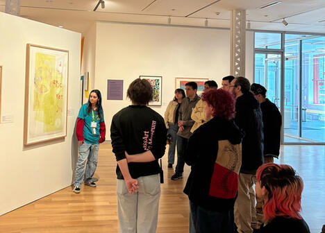 A group of people listens to a guide in front of colorful artworks in a modern gallery. The atmosphere is attentive and curious.