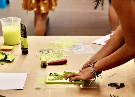 A person arranges leaves on a printing plate at a craft table with art supplies and paper nearby.