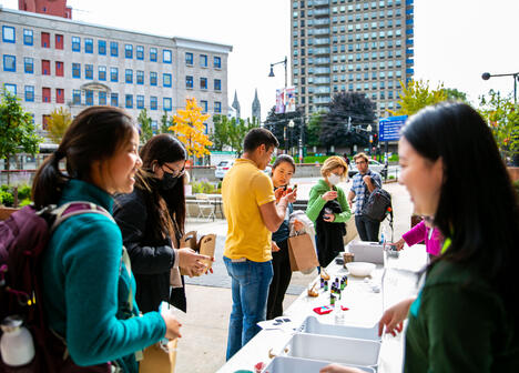 People stand in line at an outdoor table with supplies, smiling and talking, with city buildings in the background.