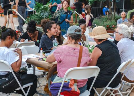 A group of people sit around a table outdoors, talking and crafting at a community event.