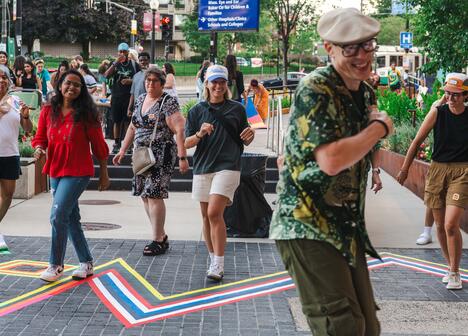 A group of people dance and smile outdoors on a city sidewalk decorated with colorful tape lines.