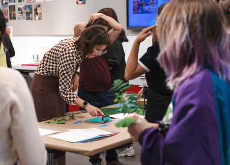 A woman demonstrates art techniques at a table while several people observe and participate in a classroom setting.