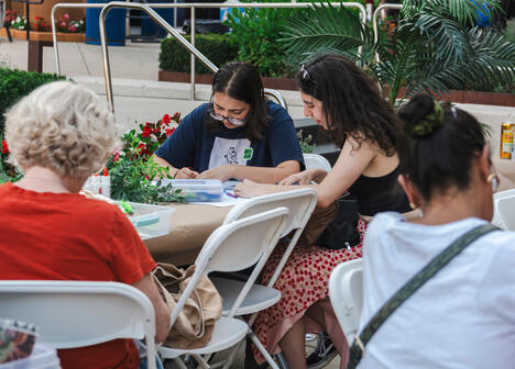 Several people sit at outdoor tables, drawing or crafting together, surrounded by plants and flowers.