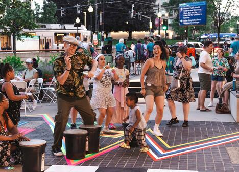 People of various ages dance and play drums outdoors at a lively community event on a city street.