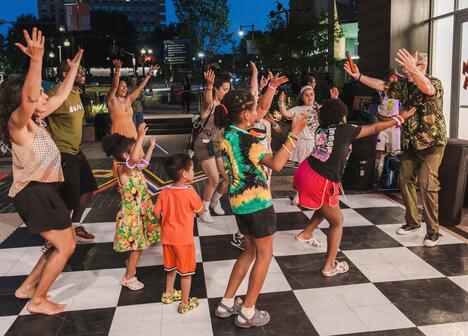 A group of adults and children dance energetically together outdoors on a black and white checkered floor at night.