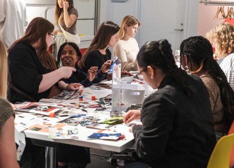 People sit around a table covered with craft supplies, talking and making art projects together indoors.