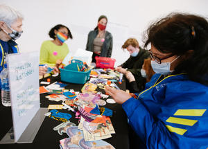 5 people of all ages seated and standing around a table covered in art making materials.