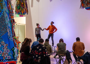 Two clothed figure models pose on a stage while visitors sketch under Joana Vasconcelos' sculpture Valkyrie Mumbet
