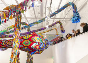 Viewers stand on a balcony in the museum looking out at the brightly colored huge sculpture hanging from the ceiling