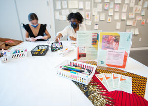 Two masked people sitting at a table with markers and cards.