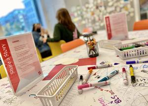 A table with markers and paper with two people in the background