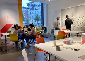 A group of people sitting and standing in a room with arts and crafts tools on a white table.