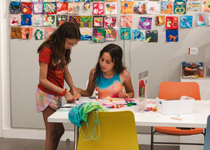 Two young girls making art at a table. One young girl is standing and the other is sitting down. Artwork made by visitors displays in the background.