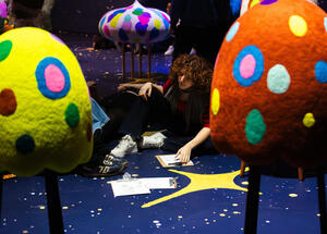 A person with curly hair sits on a starry floor, surrounded by colorful, spotted sculptures resembling mushrooms. They are drawing on a clipboard, creating a thoughtful ambiance.