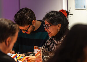 Two people sit at a table, smiling and engaged with colorful pens. The atmosphere is warm and cheerful, suggesting a creative or social activity.