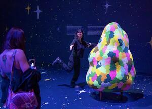 A person poses playfully beside a colorful, abstract sculpture in a dark, starry-themed room. Another person photographs with a phone, creating a joyful atmosphere.