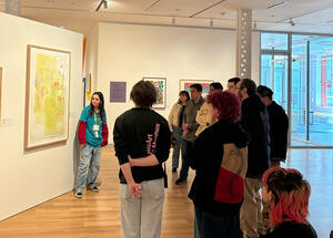 A group of people listens to a guide in front of colorful artworks in a modern gallery. The atmosphere is attentive and curious.