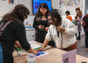 Several people work together on an arts and crafts project at a table in a brightly lit art museum space.