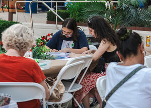 Several people sit at outdoor tables, drawing or crafting together, surrounded by plants and flowers.