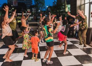A group of adults and children dance energetically together outdoors on a black and white checkered floor at night.