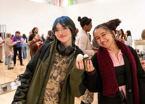 Two smiling people at an indoor event, one holding cupcakes, with a lively group in the background.