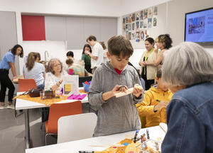 Children and adults work on art and crafts at tables in a bright, busy classroom or activity room.