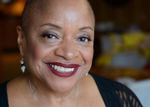 Smiling woman with short hair, wearing earrings and a beauty necklace, in a softly lit indoor setting.