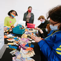 5 people of all ages seated and standing around a table covered in art making materials.
