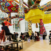 Visitors in the Paine Gallery sitting under a large colorful hanging sculpture drawing from two live models posed on a stage.