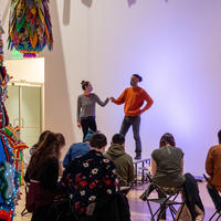 Two clothed figure models pose on a stage while visitors sketch under Joana Vasconcelos' sculpture Valkyrie Mumbet