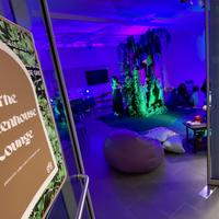 An open doorway with a sign that reads "The Greenhouse Lounge" leading into a room lit with deep purple and green lights and has low seating 