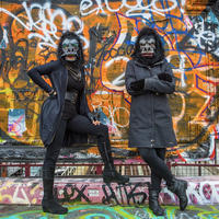 Two women dress in all black with guerrilla masks stand in front of a wall covered with grafitti 