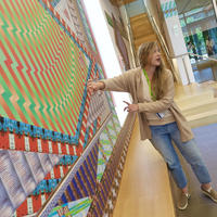 MAAM Educator with long blonde hair wearing a tan cardigan with blue jeans with her arms outstretch giving a talk about artwork on the wall 