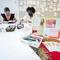 Two masked people sitting at a table with markers and cards.