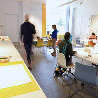 A group of people sitting and standing in a room with arts and crafts tools on a white table.
