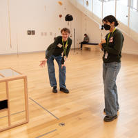 One person standing with a mask beside another person pointing at a mirrored cube.