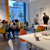 A group of people sitting and standing in a room with arts and crafts tools on a white table.