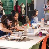 A group of people sitting at tables, engaged in arts and crafts with magazines and scissors. The setting is lively, with colorful chairs and a creative atmosphere.
