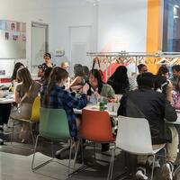 A diverse group of people engage in crafts at tables in a bright, modern room. The atmosphere is lively, with colorful chairs and creative materials.