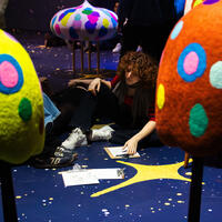 A person with curly hair sits on a starry floor, surrounded by colorful, spotted sculptures resembling mushrooms. They are drawing on a clipboard, creating a thoughtful ambiance.