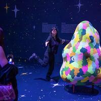A person poses playfully beside a colorful, abstract sculpture in a dark, starry-themed room. Another person photographs with a phone, creating a joyful atmosphere.