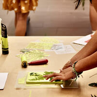 A person arranges leaves on a printing plate at a craft table with art supplies and paper nearby.