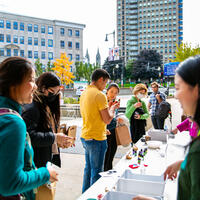 People stand in line at an outdoor table with supplies, smiling and talking, with city buildings in the background.